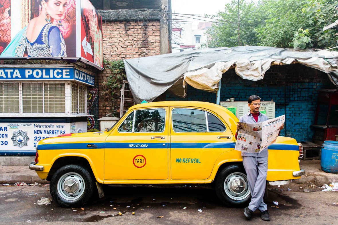 A man reads a newspaper in front of a classic yellow Ambassador taxi cab in Kolkata, West Bengal, India.
